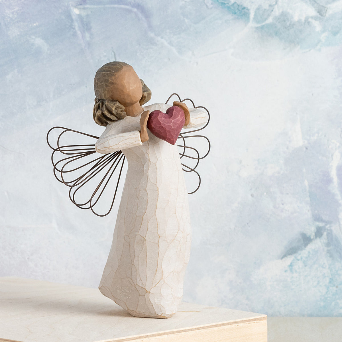 Willow Tree With Love, 5.5”h hand-painted standing resin angel figure with darker skin and hair, in cream dress with wire wings, holding a red heart in hands, in front of blue painting
