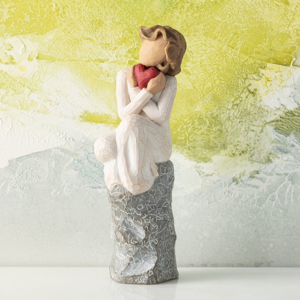 Willow Tree Always, 6”h hand-painted figure in cream dress, knees drawn up to chest, holding red heart to face, seated on gray rock etched with symbols of love, in front of lime painting