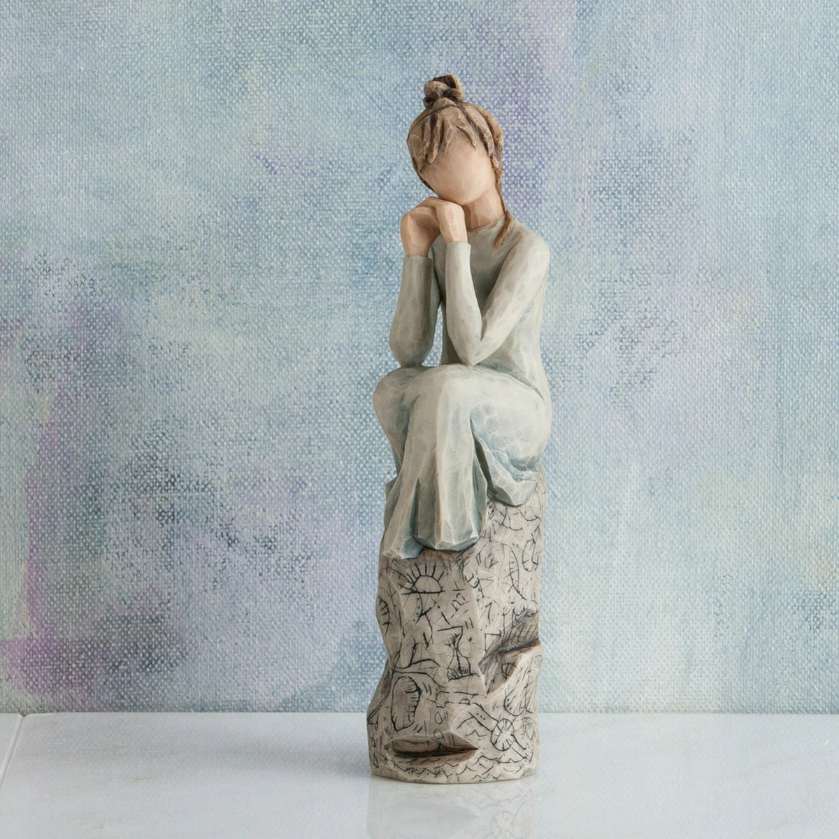 Willow Tree Patience, 7”h hand-painted resin figure in blue/gray dress, hands under chin, sitting on grey rock etched with symbols, in front of muted blue painting