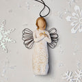 Willow Tree Remembrance Ornament, 4.5”h hand-painted resin angel figure in cream dress carved with symbols of nature, wire wings, crossed hands under small gold-leaf heart, brown elastic cord, laying amongst snowflakes