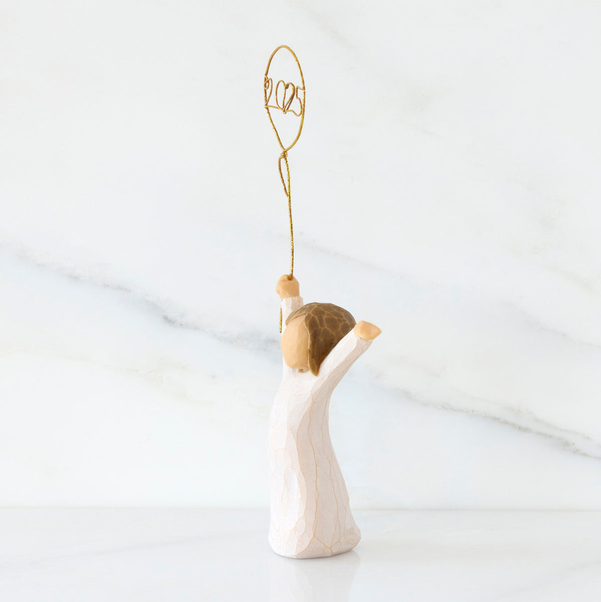 Facing left: standing figure in cream dress, arms raised, holding gold wire balloon with 2025 written in gold wire inside