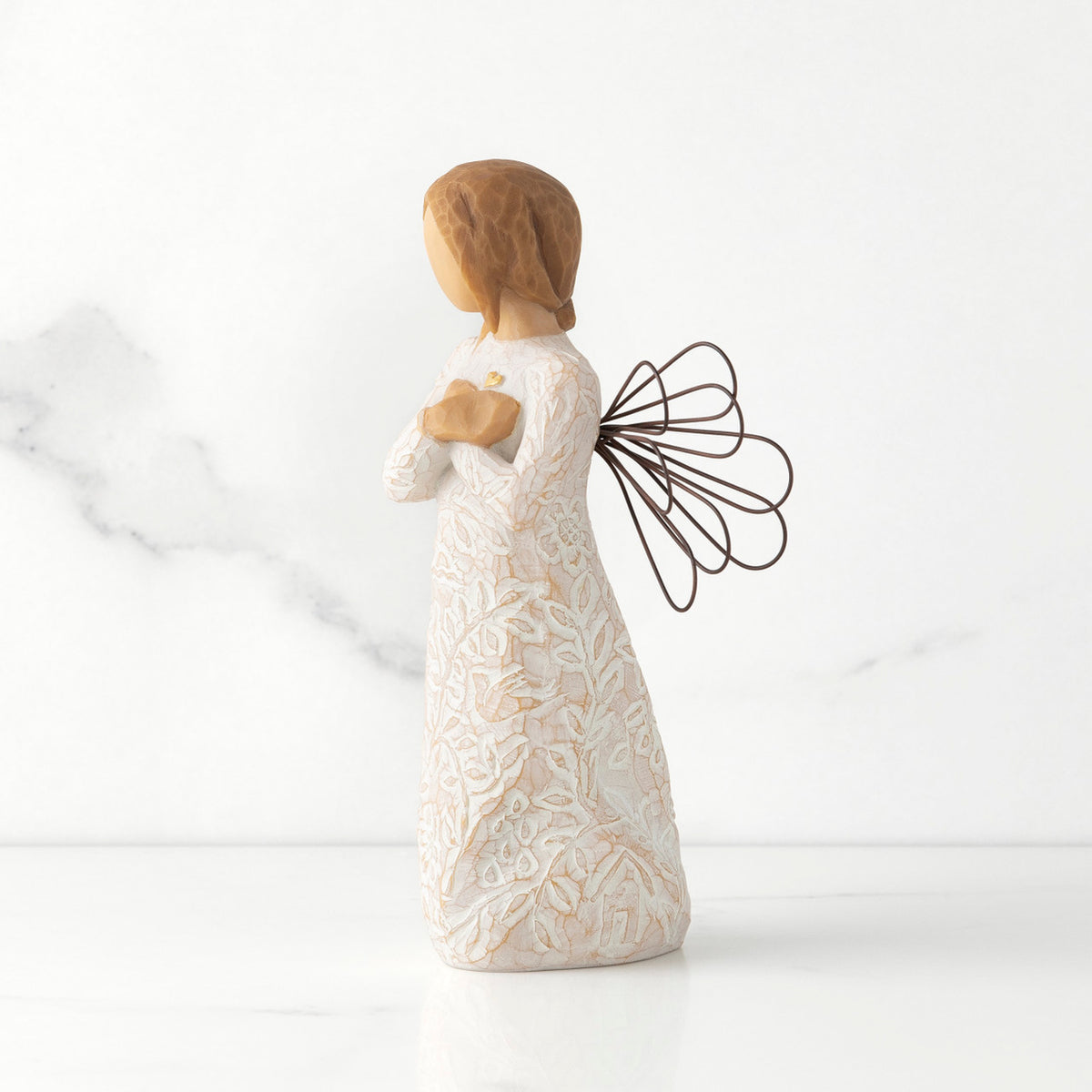 Left side view of standing female figure with wire wings in cream dress carved with natural symbols, hands crossed under gold heart on chest