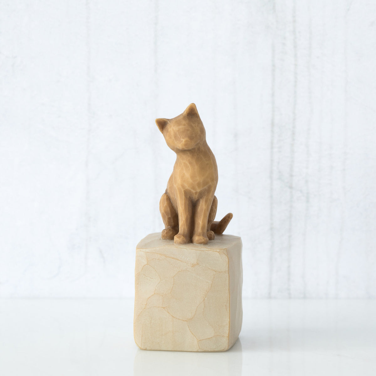 Figure of orange cat with tilted head perched atop a cream colored rectangular pedestal, facing front