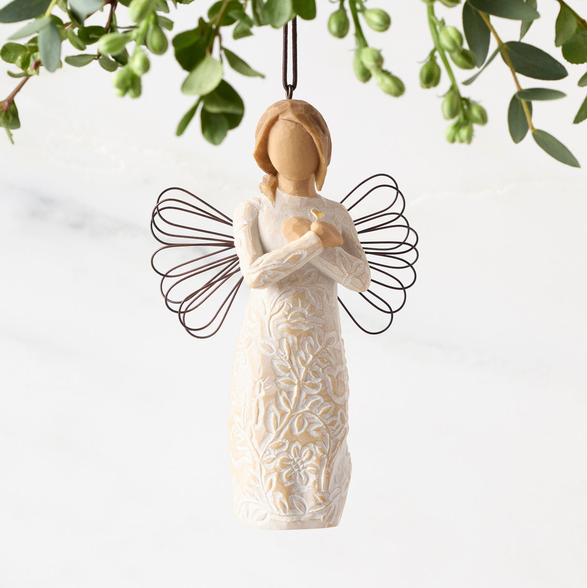 Front view of figure in cream dress carved with symbols of nature with wire wings, hands folded at chest around gold heart, ornament hook on head