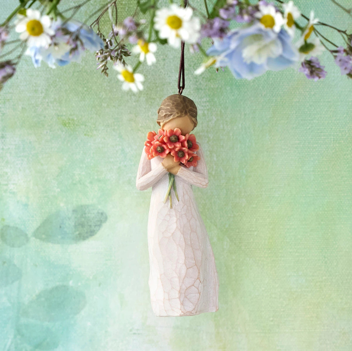 Willow Tree Surrounded by Love Ornament, 4”h hand-painted resin figure in cream dress holding bouquet of poppies to face, brown elastic cord, hanging in front of pale green painting