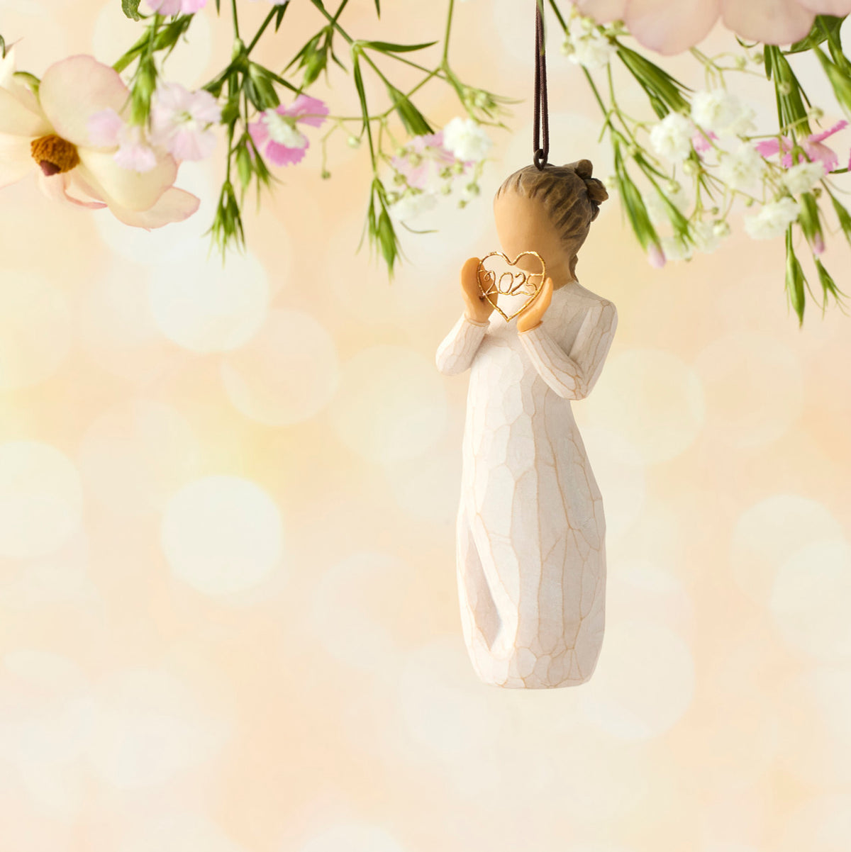 Willow Tree 2025 Ornament, 4”h hand-painted resin figure in cream dress, holding wire heart with 2025 inside, brown elastic cord, hanging in front of peach painting