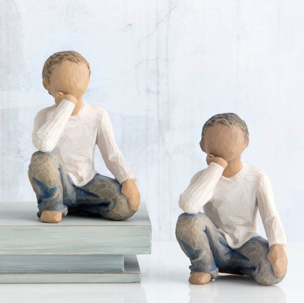 Two young brunette boy figurines in white and jeans sitting down with one arm to their face next to stack of books