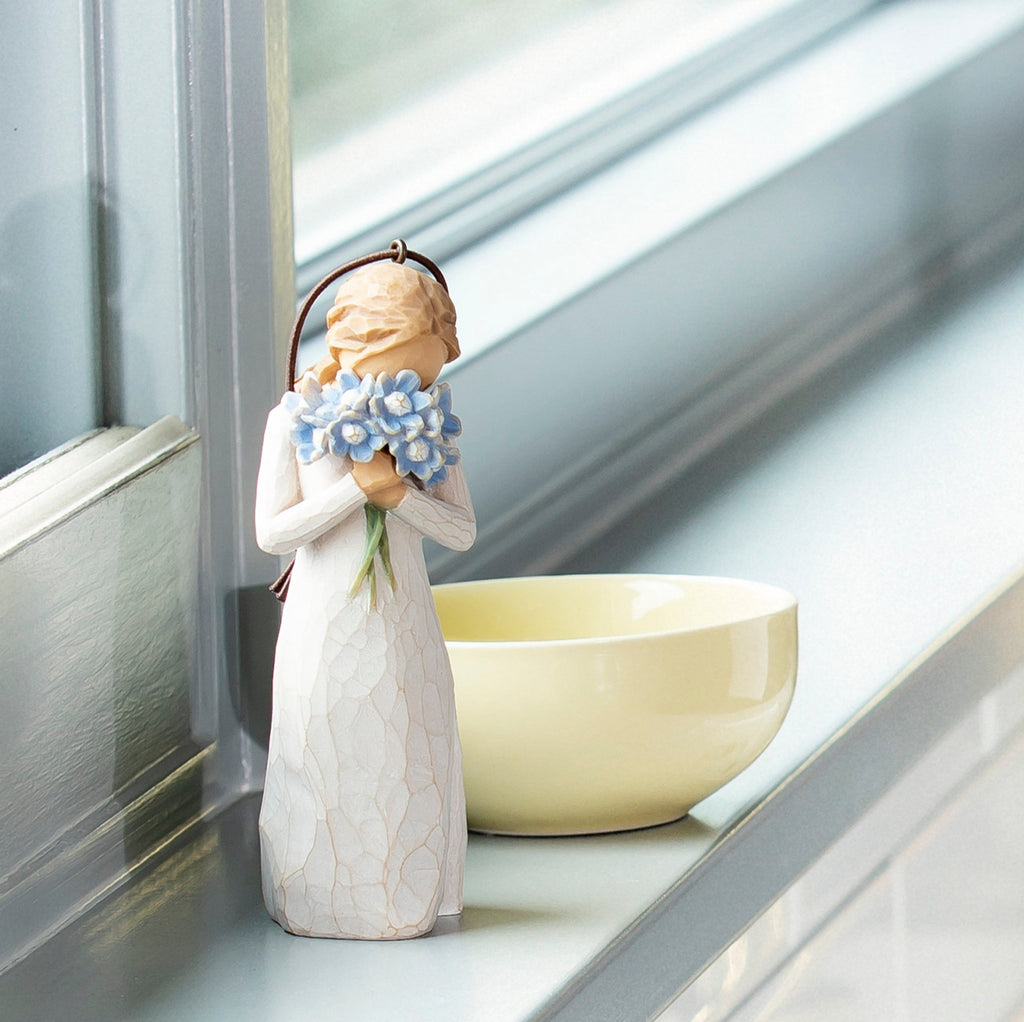 Willow Tree Forget-me-not Ornament, 4.25”h hand-painted resin figure in cream dress, holding bouquet blue flowers up to face, brown elastic cord, on bright windowsil