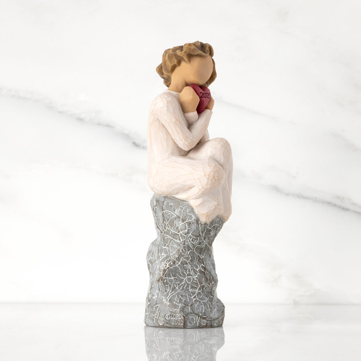 Facing right: figure in cream dress, seated on gray rock, knees drawn up to chest, holding red heart engraved with year 2023