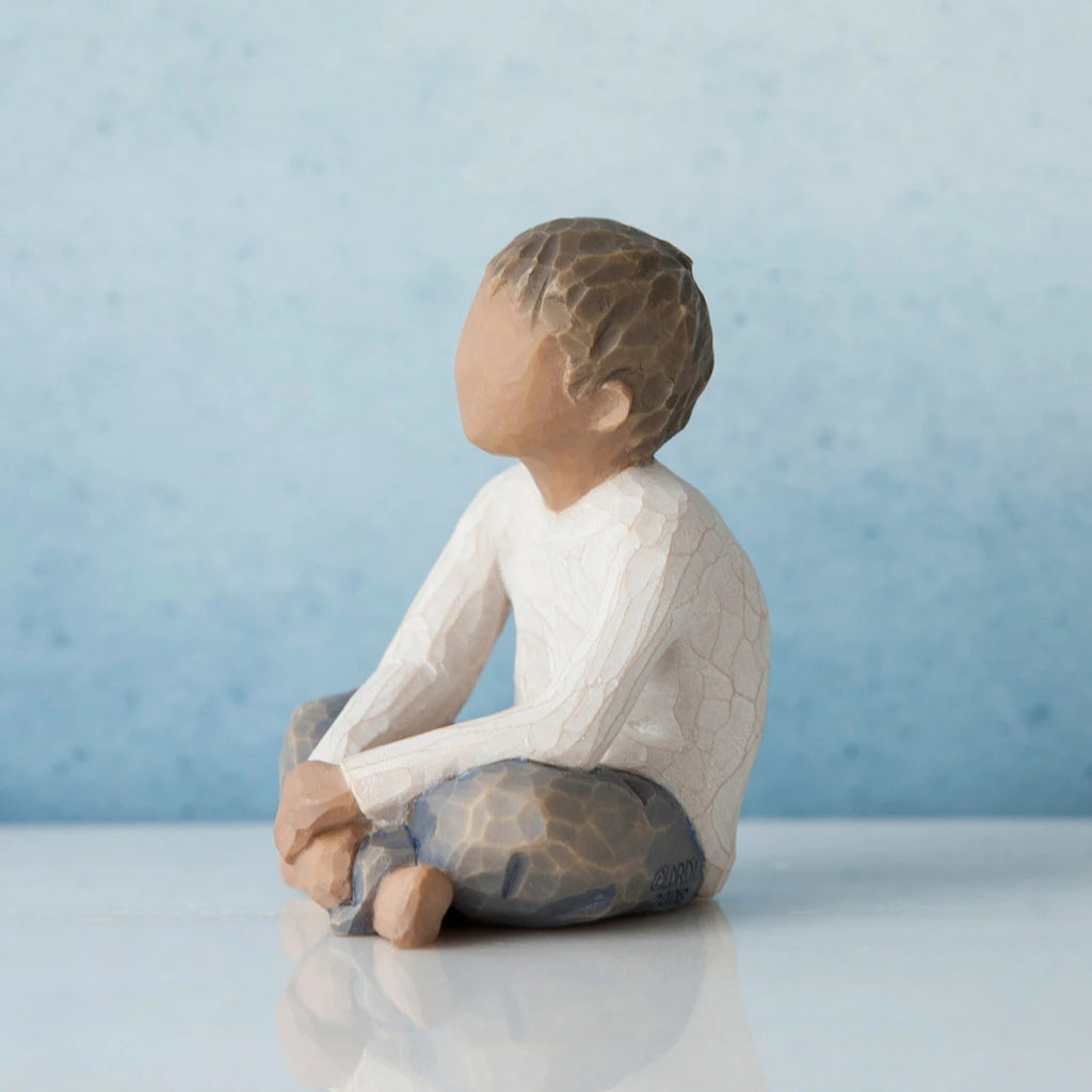 Small brunette boy figurine sitting to the side wearing white shirt and blue jeans and hands in lap - blue background