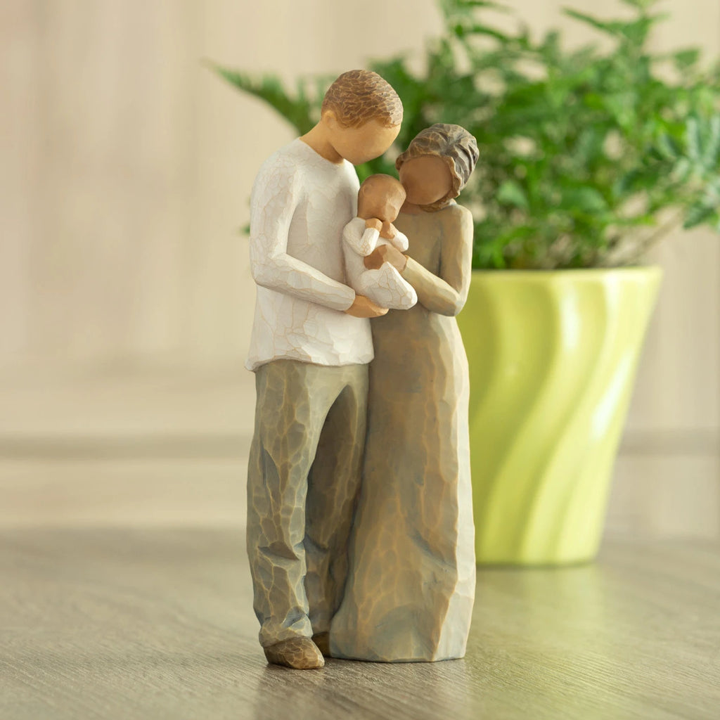 Front view of standing couple, woman has darker skin and hair; man has lighter skin and hair, holding a baby between them. Potted plant in background.