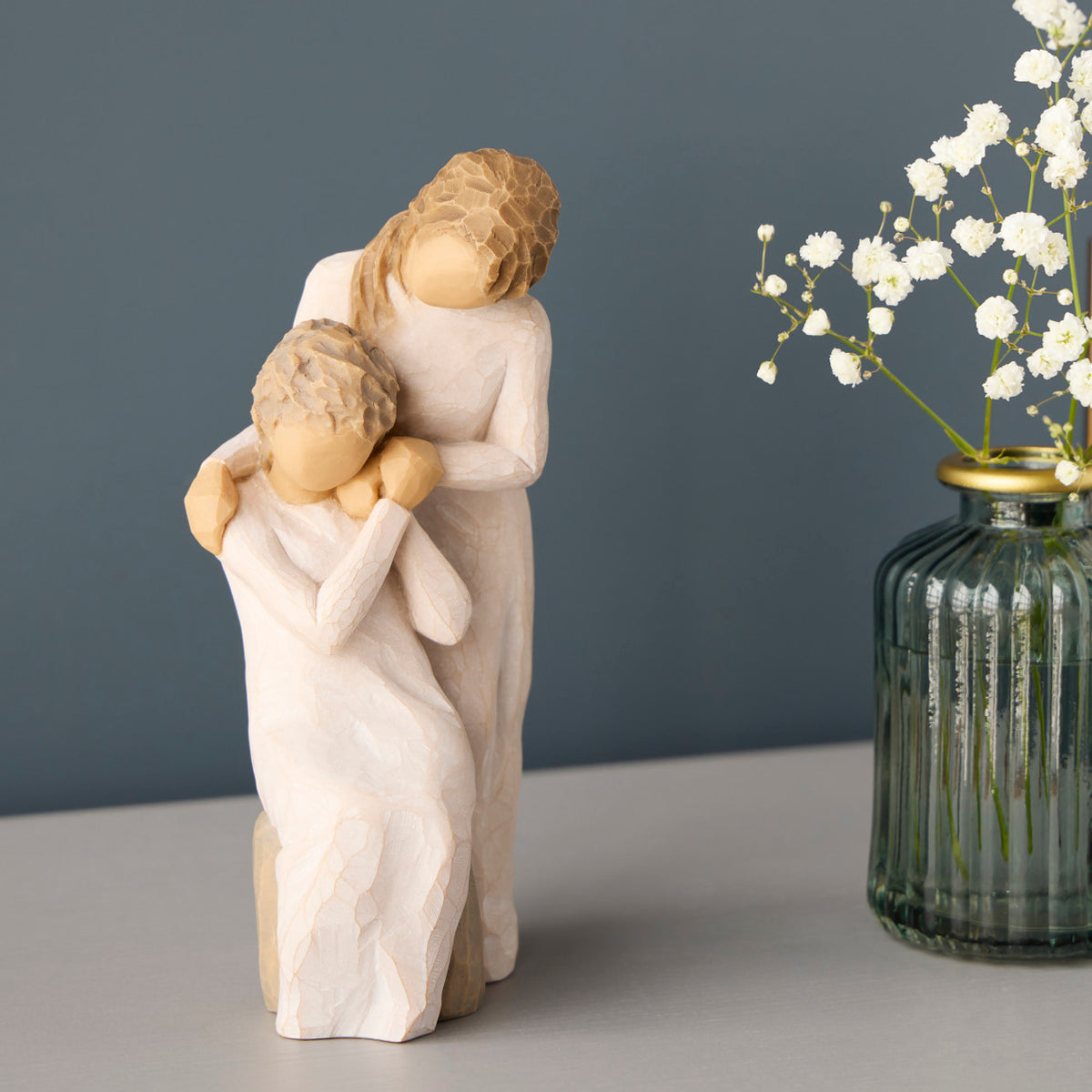 Willow Tree Loving My Mother, 6.5”h hand-painted resin figure of seated older woman with younger woman standing at her shoulder, both in cream dresses, hands clasped, on table with white flowers
