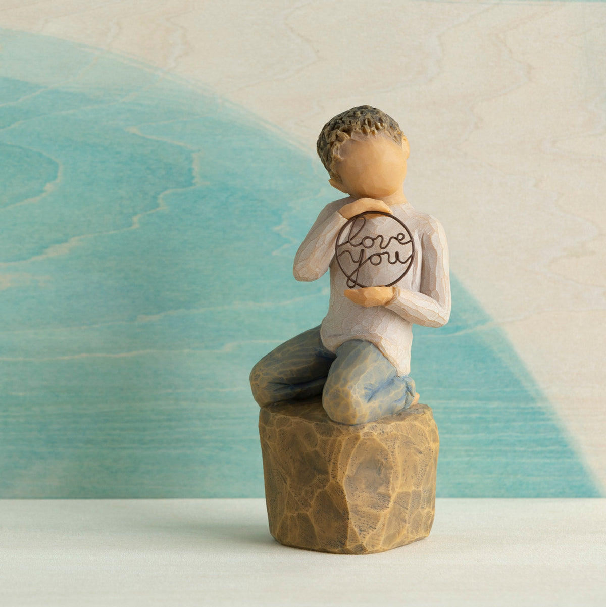 Willow Tree Love you too (lighter skin), 4.5”h hand-painted resin figure of young child in cream shirt and jeans, kneeling on rock, holding wire circle with ‘love you’ inside, in front of blue painting