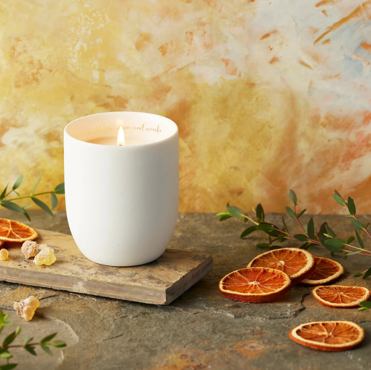 Willow Tree Festive Orange and Frankincense Candle, 3.5"h off-white ceramic vessel with plant-based wax and blend of natural oils, "behold the awe and wonder" sentiment in gold inside rim, in front of orange painting